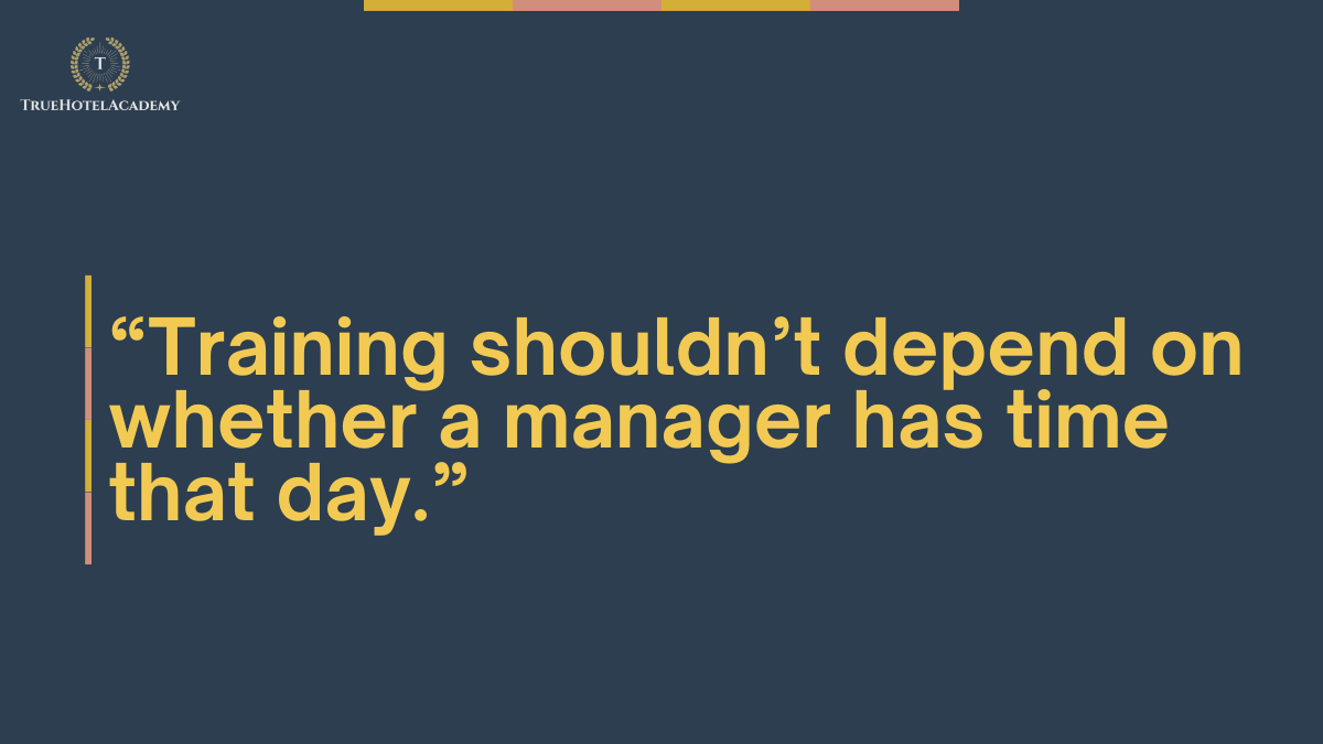 A dark blue background with the TrueHotelAcademy logo in the top left corner. The logo features a stylized 'T' within a wreath. A quote in bold yellow text reads: 'Training shouldn't depend on whether a manager has time that day.' The quote is framed by vertical yellow and pink lines on the left. Thin horizontal stripes in yellow, light pink, and dark pink are visible at the very top of the image.
