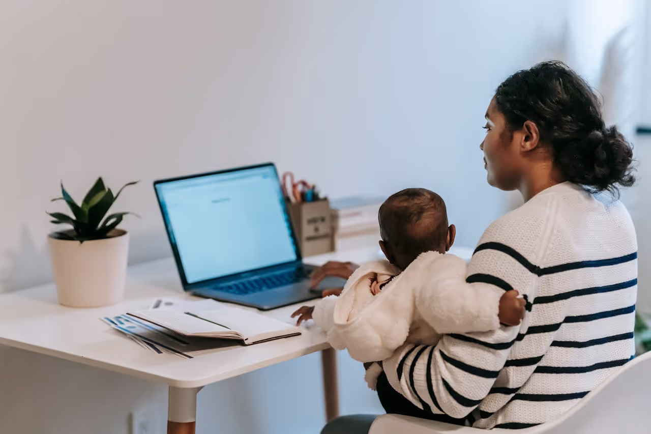 Student mother working on school assignments in front of a laptop while holding her baby, showing how colleges support student parents college success.