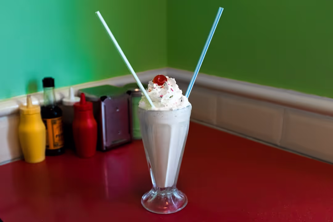 A tall glass of creamy vanilla milkshake topped with whipped cream and a red cherry.