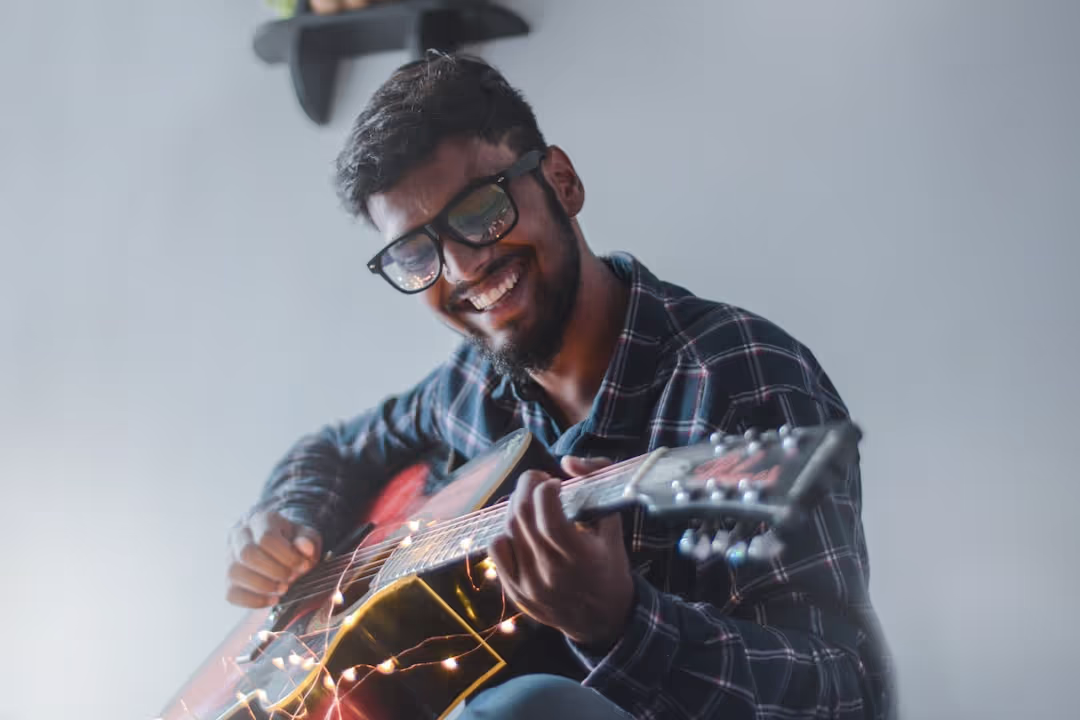 A smiling young man with glasses and a beard plays an acoustic guitar decorated with fairy lights, looking happily at the viewer.