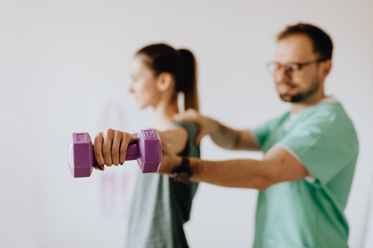 Side view of professional orthopedist in uniform and eyewear helping fit woman reaching arm with dumbbell in doctor office on blurred background 4506112